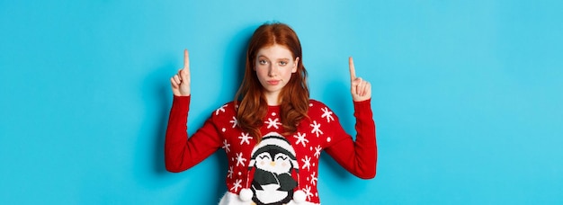Free photo merry christmas serious and confident redhead girl pointing fingers up showing advertisement and sta