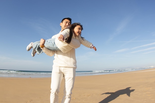 무료 사진 메리 아버지와 딸이 해변에서 놀고 있습니다. 화창한 날 일본 가족들이 비행기를 모방하고 웃고 있습니다. 아빠 여가, 가족 시간, 육아 개념