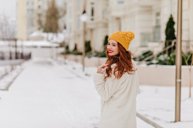 冬の街を歩きながら振り返る陽気な生姜の女性。雪の朝に身も凍る優雅なヨーロッパの女の子。