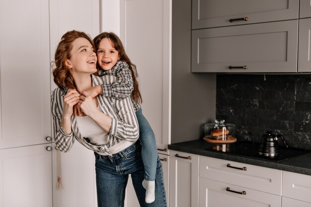 Merry mom and daughter in similar outfits play in kitchen and laugh.