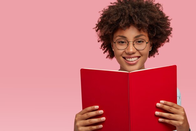 Merry pleased African American woman with pleased expression, wears optical glasses for good vision, holds textbook, learns material for seminar, isolated over pink wall with blank space on left