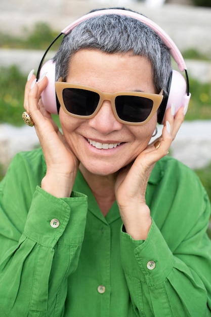 Бесплатное фото Веселая пожилая женщина слушает музыку в парке. женская модель с короткими седыми волосами в яркой одежде и большими наушниками. досуг, активность, технологическая концепция