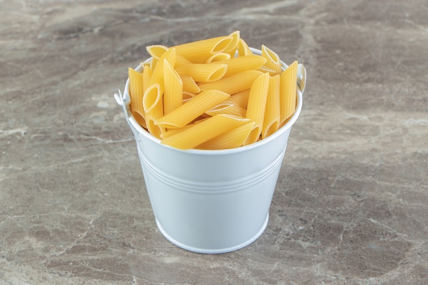 Metal bucket of raw penne on blue surface