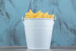 Free photo metal bucket of raw penne on blue surface.