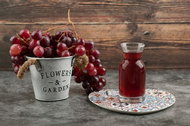 Металлическое ведро красного свежего винограда и стакан сока на мраморном столе.