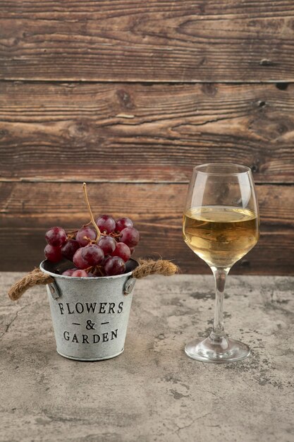Металлическое ведро красного свежего винограда и бокал белого вина на мраморной поверхности.