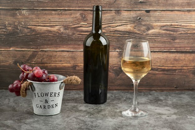 Metal bucket of red fresh grapes with bottle of white wine on marble table.