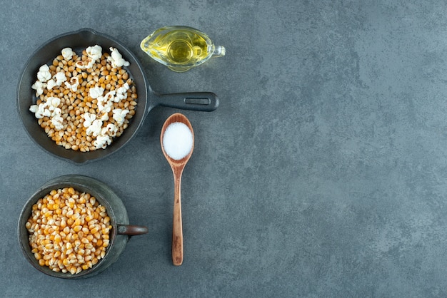 Free photo metal jug and pan filled with corn grains, with a spoonful of salt and a glass of oil on marble background. high quality photo