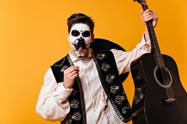 Mexican brown-eyed man with face art in form of skull emotionally shouts, posing with false mustaches and guitar in his hands on orange wall.