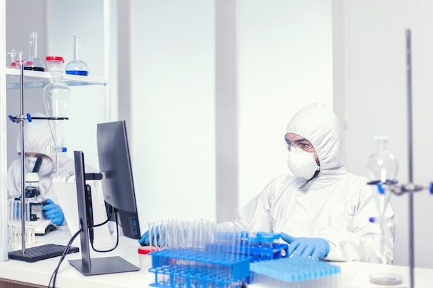 Microbiologist dressed in sterile ppe suit researching for covid vaccine looking at pc screen. Medical engineer using computer during global pandemic with coronavirus dressed in coverall.