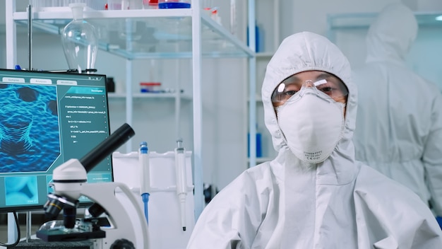 실험실에 앉아 있는 미생물학자는 현대적인 장비를 갖춘 실험실에서 카메라를 보고 있는 PPE 정장을 입고 있습니다. 과학 연구를 위한 첨단 화학 도구를 사용하여 바이러스 진화를 조사하는 과학자 팀