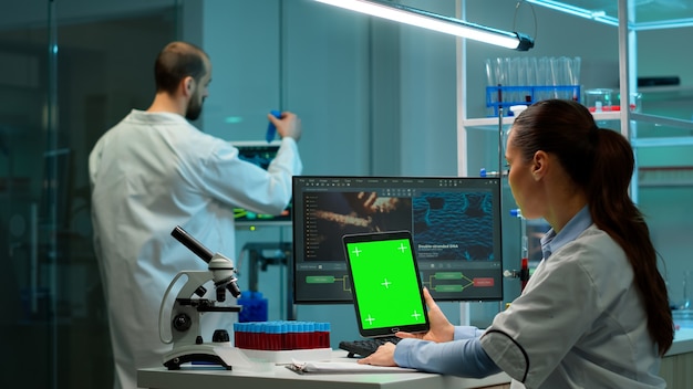 Microbiologist working on notepad with green chroma key display in modern equipped lab. Team of biotechnology scientists developing drugs using tablet with mock up screen.