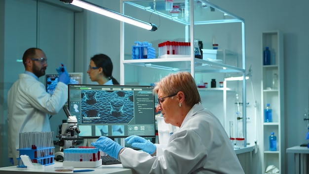 Microchemist doing research at night checking blood liquid in test tube typing results on pc in modern equipped lab. Examining virus evolution using high tech for vaccine development against covid19