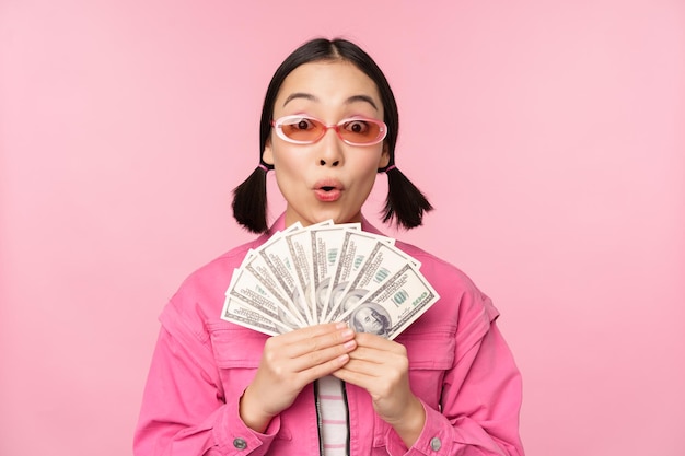 Microcredit and fast loans concept Excited stylish korean girl showing money cash dollars and looking happy standing in sunglasses over pink background
