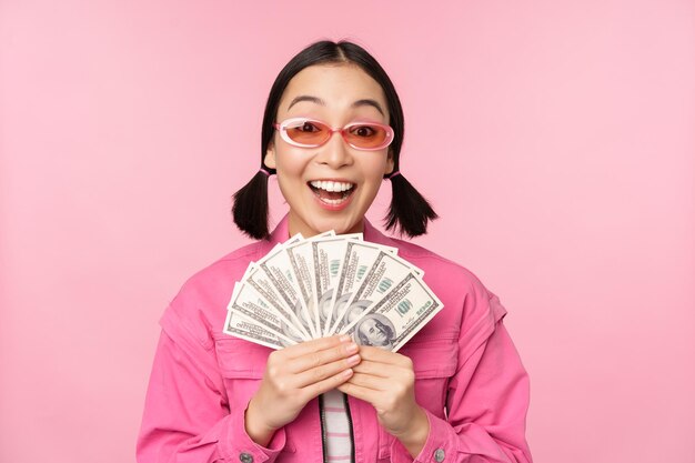 Microcredit and fast loans concept Excited stylish korean girl showing money cash dollars and looking happy standing in sunglasses over pink background