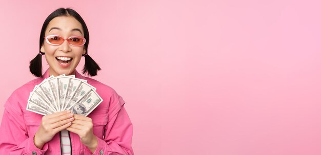 소액 신용 및 빠른 대출 개념 현금 달러를 보여주고 분홍색 배경 위에 선글라스를 끼고 행복하게 서 있는 것처럼 보이는 흥분된 세련된 한국 소녀
