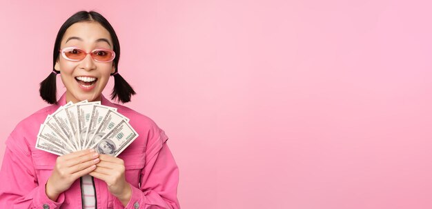 Концепция микрокредитов и быстрых кредитов Взволнованная стильная корейская девушка показывает деньги наличными долларами и выглядит счастливой, стоя в солнцезащитных очках на розовом фоне