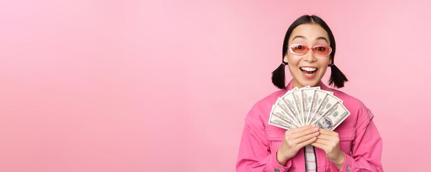 Microcredit and fast loans concept Excited stylish korean girl showing money cash dollars and looking happy standing in sunglasses over pink background