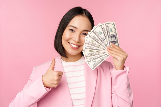 Free Photo microcredit investment and business people concept young asian businesswoman corporate lady showing money cash dollars thumbs up recommending company pink background