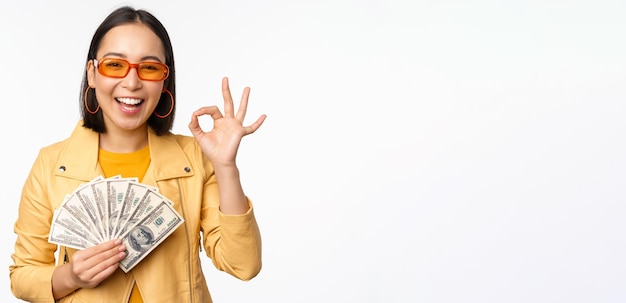 Концепция микрокредитования и кредита счастливая стильная корейская девушка показывает хорошо знак ок и деньги доллары наличными