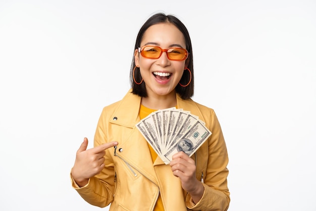 Free photo microcredit and money concept stylish asian young woman in sunglasses laughing happy holding dollars cash standing over white background
