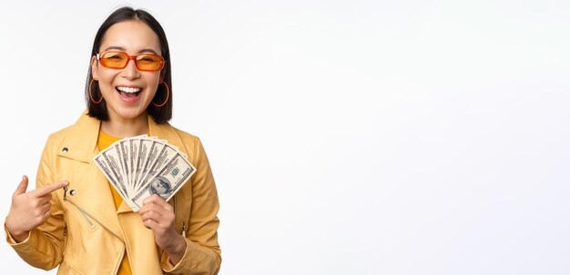 Microcredit and money concept Stylish asian young woman in sunglasses laughing happy holding dollars cash standing over white background