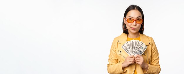 소액 신용 및 돈 개념 선글라스를 쓴 세련된 아시아 젊은 여성이 달러를 들고 행복하게 웃고 있습니다.