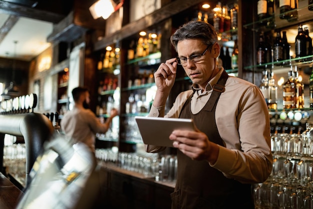 Бариста среднего возраста использует цифровой планшет во время работы в баре. Его коллега на заднем плане