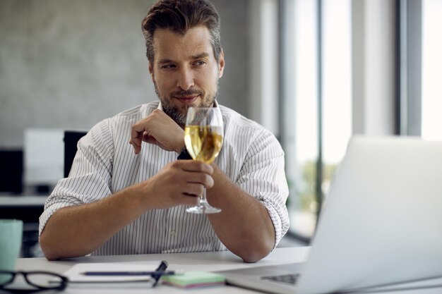 Mid adult businessman toasting with glass of wine while having video call over laptop in the office