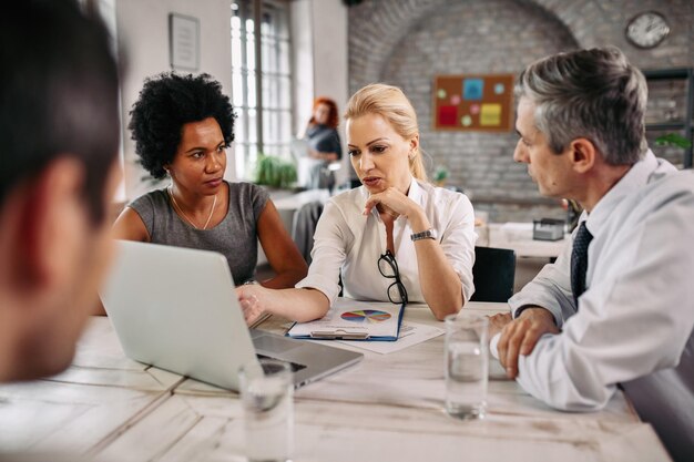 Mid adult businesswoman talking to her colleagues and while using laptop and pointing on the screen in the office