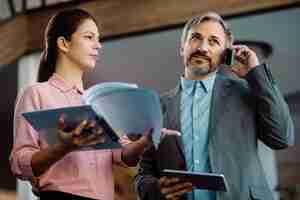 Free photo mid adult ceo making a phone call while female colleague is showing him business reports in a hallway