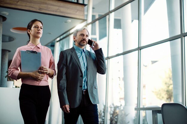 Mid adult CEO talking on the phone while walking through a hallway with female colleague