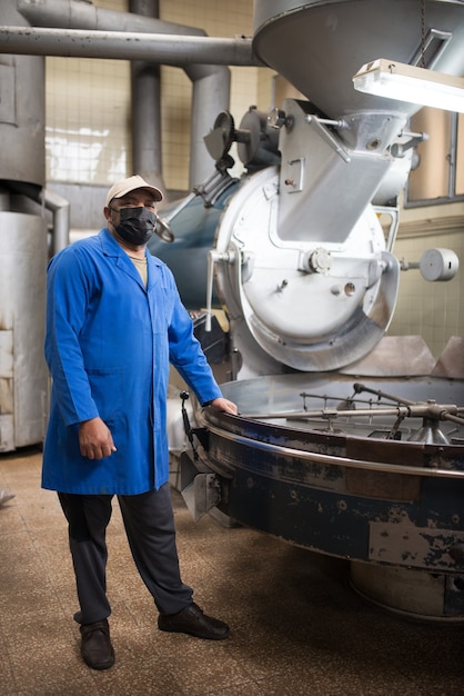 Free photo mid adult coffee roaster standing next to coffee roasting machine. interior of coffee production workshop with set up roasting equipment