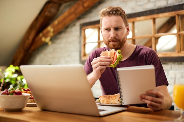 Mid adult man eating sandwich while using laptop at dining table