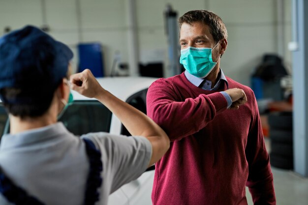 Mid adult man and female mechanic greeting with elbow while wearing protective face masks in auto repair shop