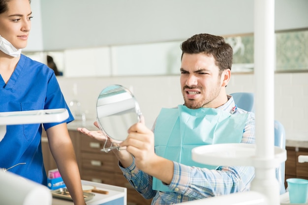 Мужчина среднего возраста с болью смотрит в зеркало, жестикулируя на женщину-стоматолога в клинике