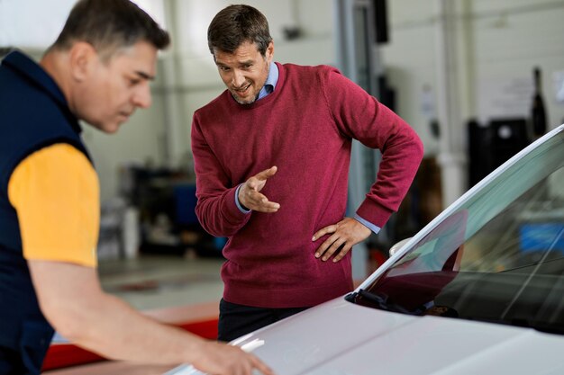 Mid adult man pointing at vehicle hood while talking to car mechanic in auto repair shop