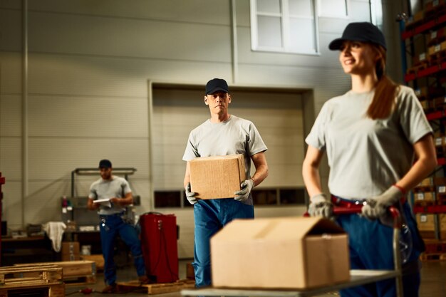 Mid adult worker carrying cardboard box while working with colleagues in industrial warehouse