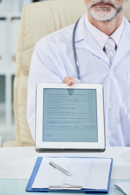Средняя часть мужчины-врача, сидящего за столом и распространяющего цифровую анкету на экране планшета на камеру