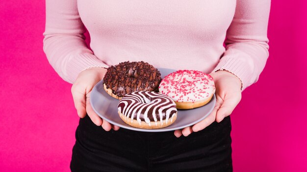 Mid section of a woman holding plate of various tasty donuts in hand against pink background