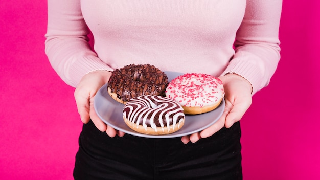 Средняя часть женщины, держащей тарелку различных вкусных пончиков в руке на розовом фоне