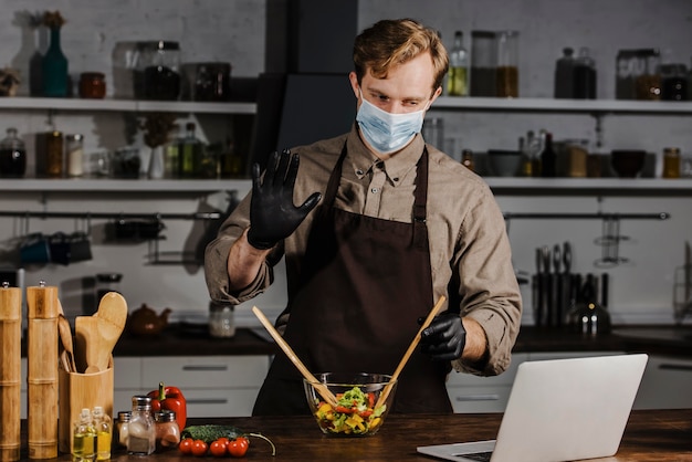 Mid shot chef with mask mixing salad ingredients looking at laptop