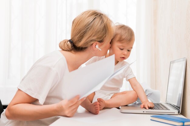 Mid shot mother holding papers and kissing baby on desk