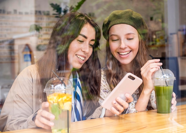 Mid shot women with fresh juices in cafe looking at phone