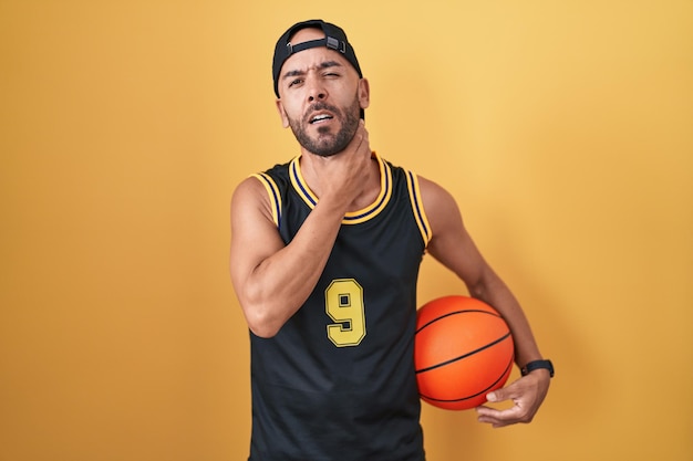 Лысый мужчина средних лет держит баскетбольный мяч на желтом фоне, касаясь болезненной шеи, боли в горле от гриппа, кома и инфекции