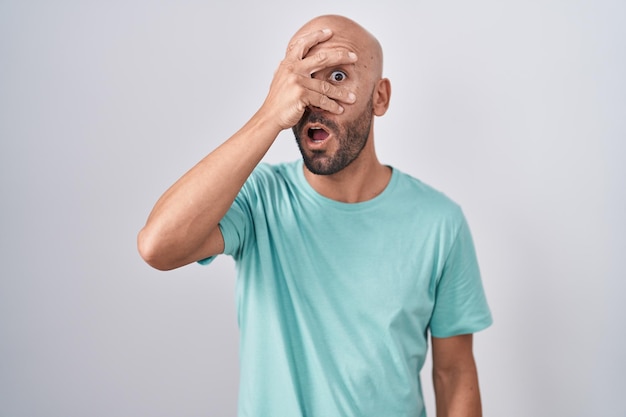 Free photo middle age bald man standing over white background peeking in shock covering face and eyes with hand, looking through fingers with embarrassed expression.