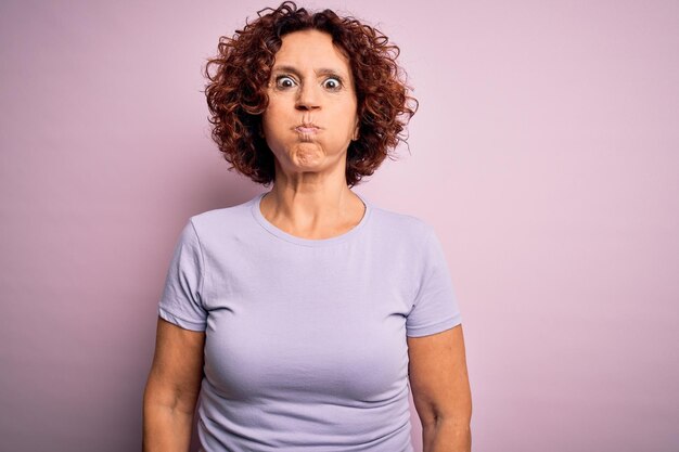 Middle age beautiful curly hair woman wearing casual tshirt over isolated pink background puffing cheeks with funny face Mouth inflated with air crazy expression