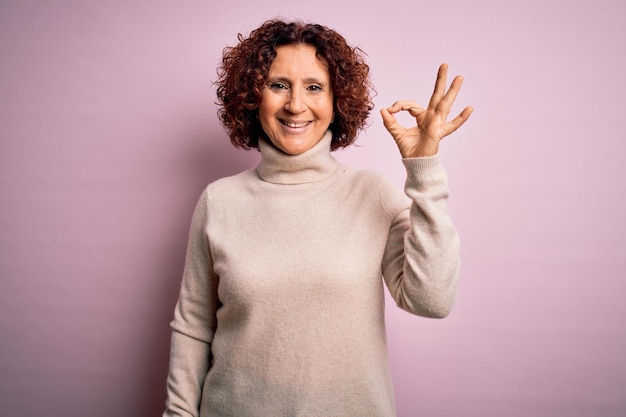 Foto gratuita donna di mezza età bella capelli ricci che indossa un maglione a collo alto casual su sfondo rosa sorridente positivo facendo segno ok con la mano e le dita espressione di successo