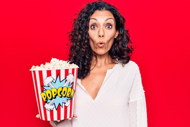 Foto gratuita bella donna di mezza età che mangia popcorn spaventata e stupita con la bocca aperta per sorpresa, faccia incredula