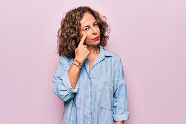 Middle age beautiful woman wearing casual denim shirt standing over pink background Pointing to the eye watching you gesture suspicious expression